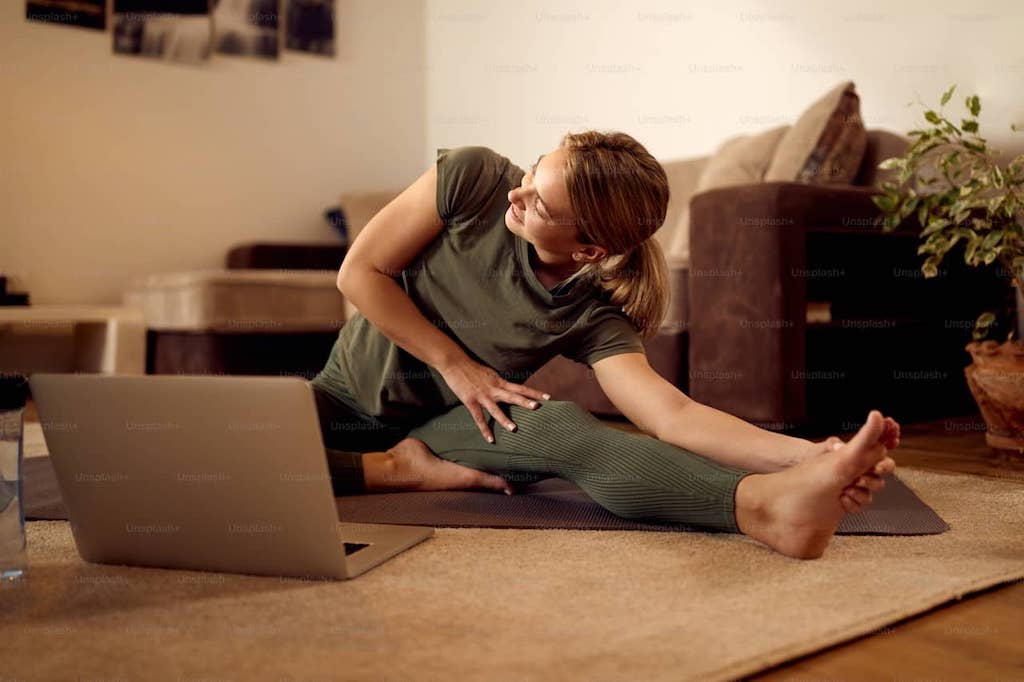 A woman manages stress by doing yoga at home, wearing all green and stretching while watching the instructor on her laptop.